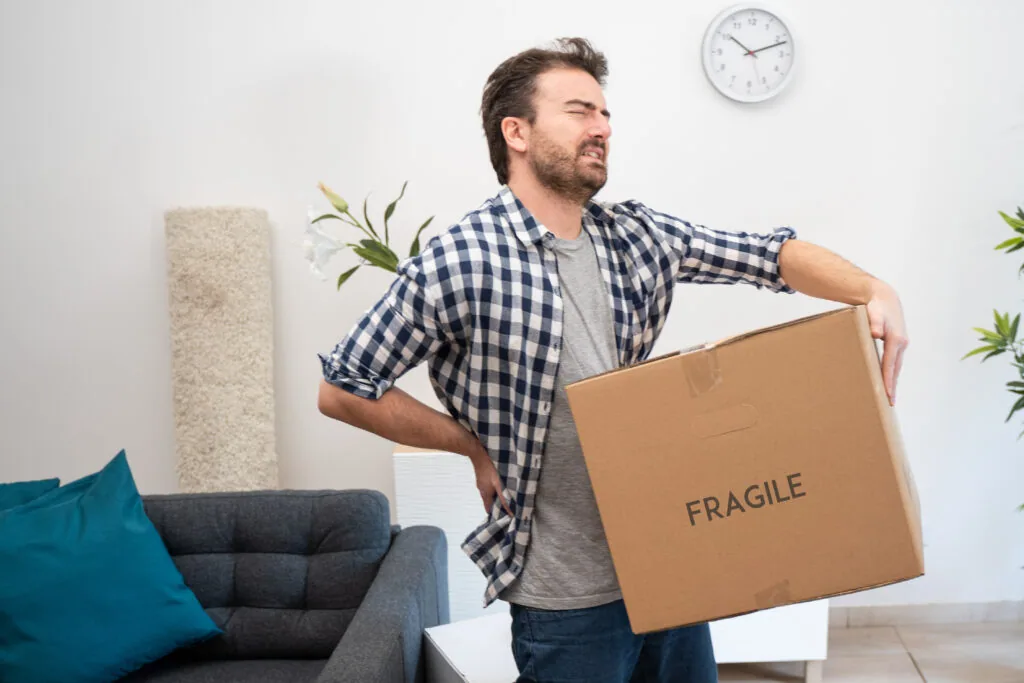 Man in checkered shirt carrying cardboard box labeled FRAGILE in living room with furniture and wall clock