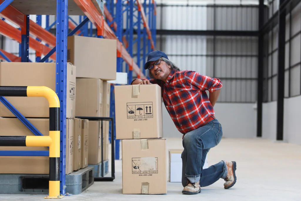 Worker in plaid shirt and cap stacking cardboard boxes in warehouse with blue metal shelving