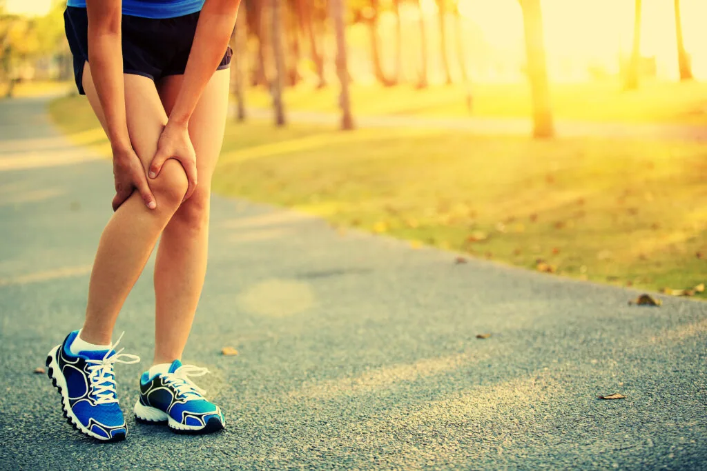 Runner holding knee in pain while standing on outdoor path at sunset