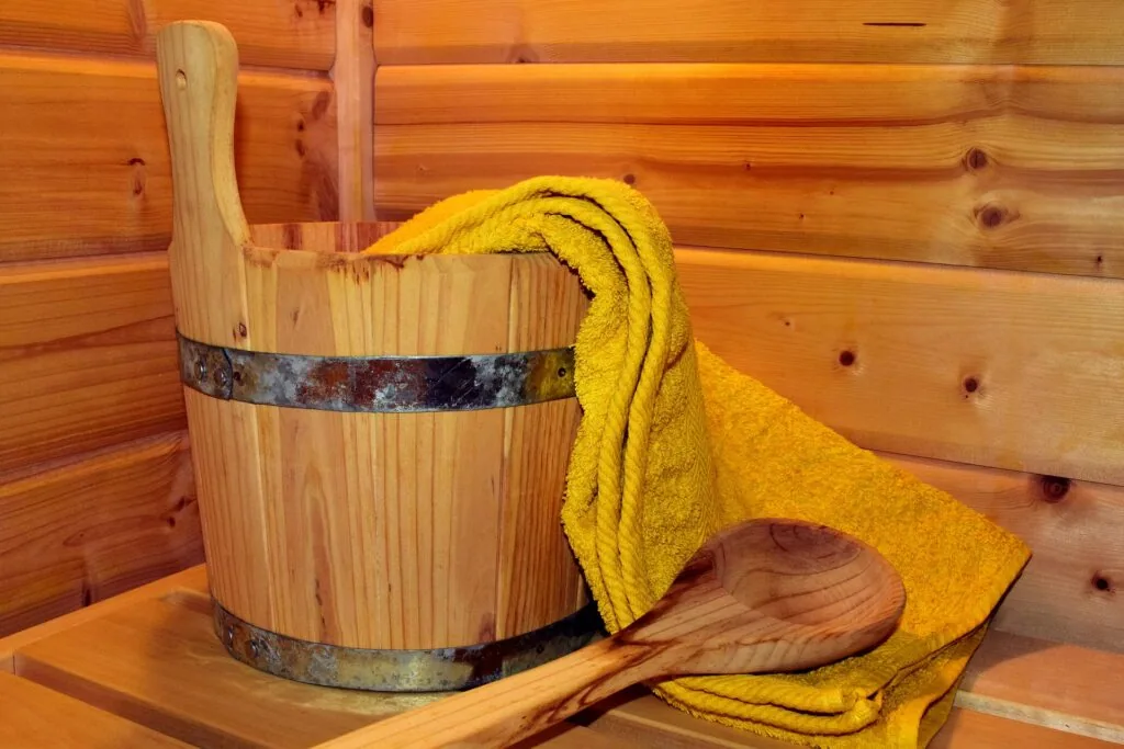 Wooden sauna bucket with metal bands, wooden ladle, and yellow towel draped over the side, set against log cabin walls